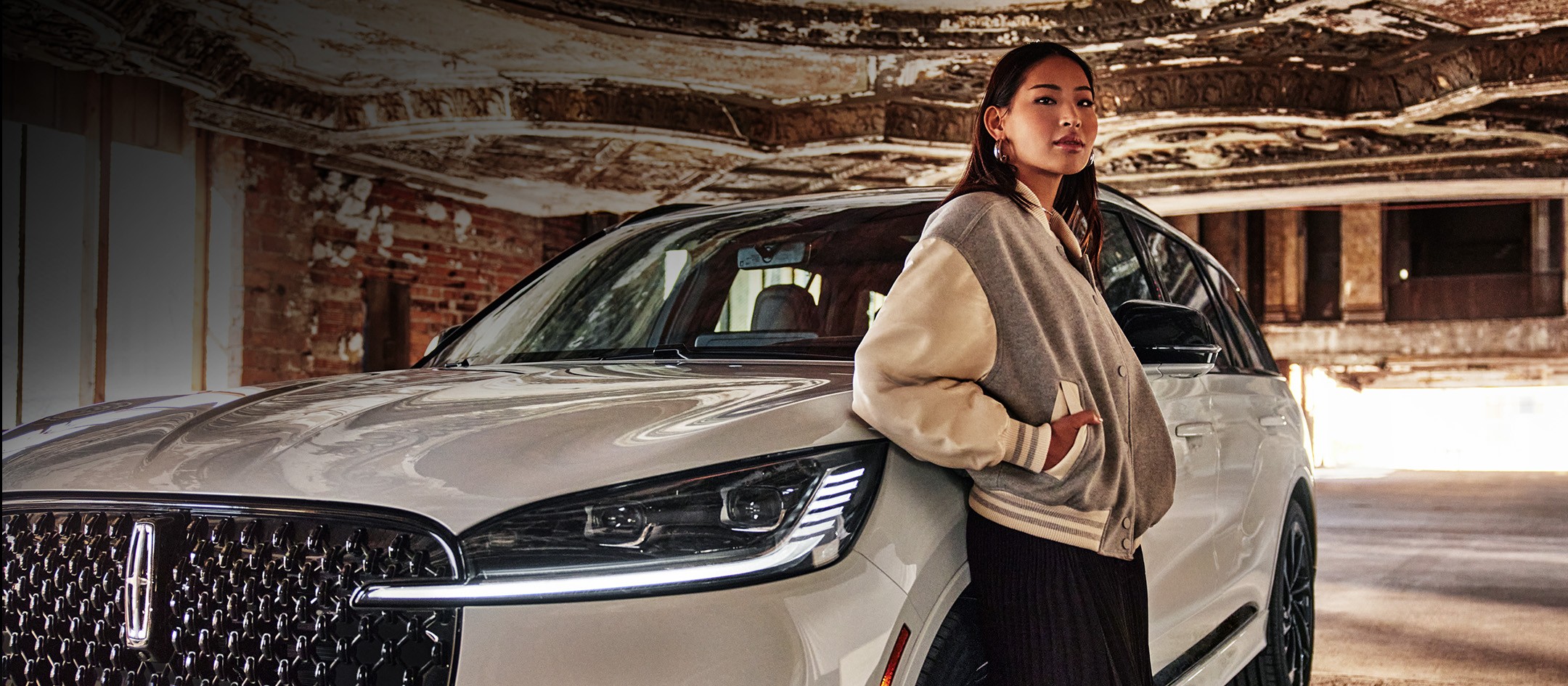 Woman in a jacket stands confidently beside a luxury SUV in an urban setting.