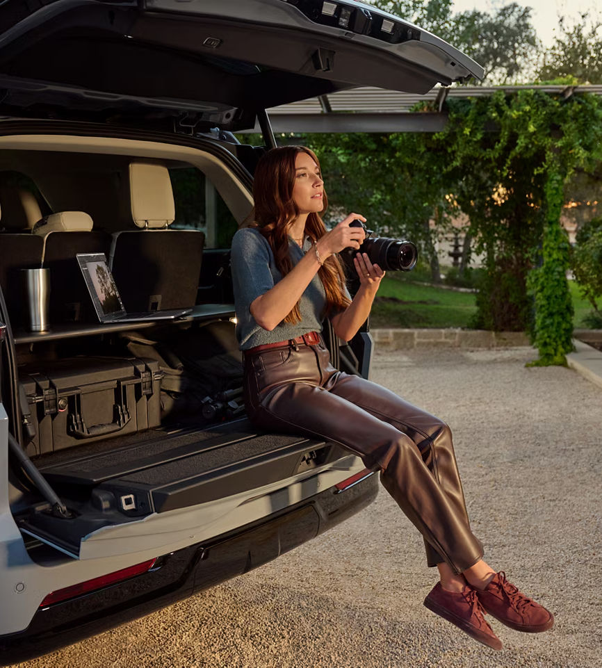 Woman sitting in SUV trunk, holding a camera, outdoors. Photography, travel, adventure scene.