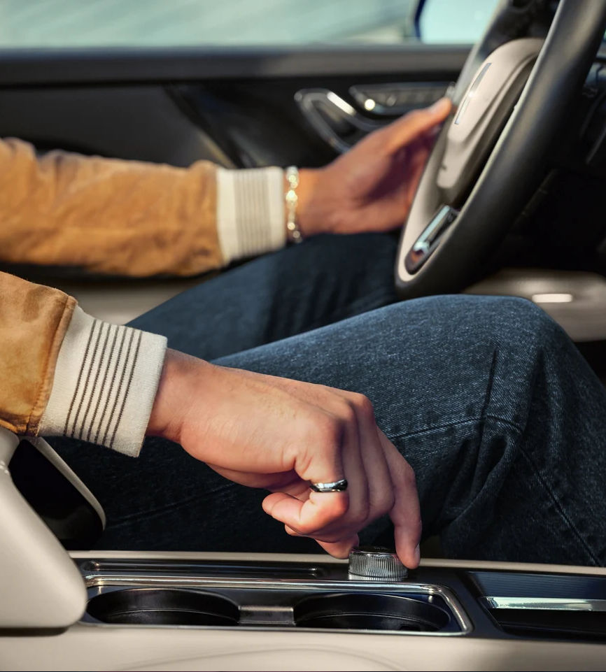 Man adjusting car controls while driving, wearing a suede jacket and ring, focusing on luxury interior details.