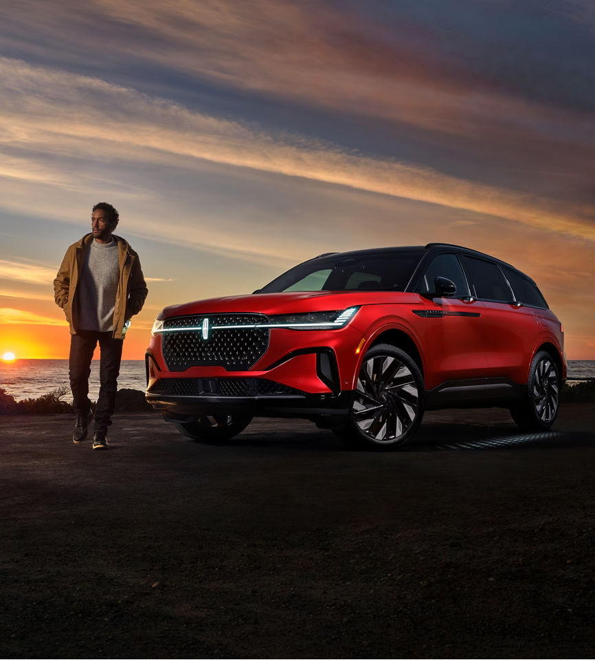 Red SUV near ocean at sunset with man in jacket, showcasing luxury automotive design.