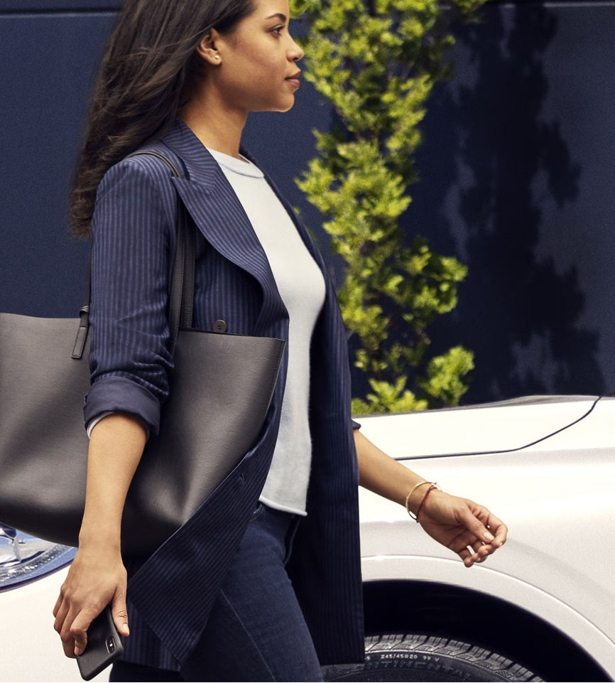 Woman in a pinstripe blazer walking with a large black tote, holding a smartphone, near a white car.