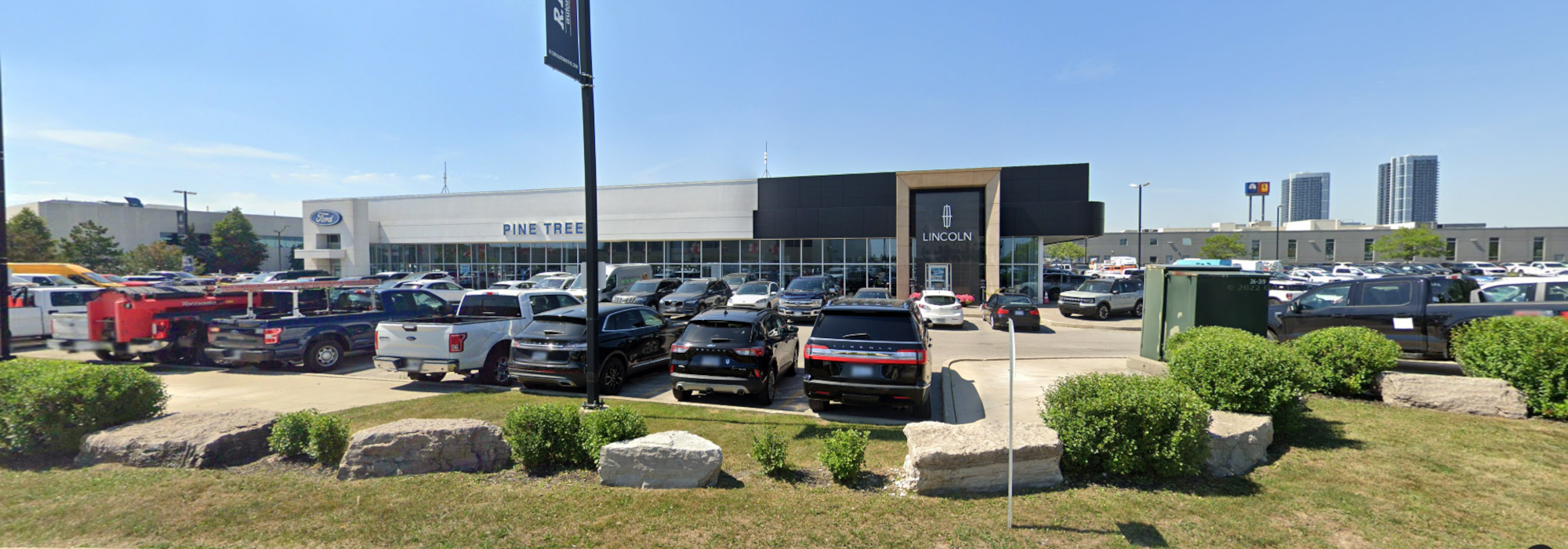 Car dealership with Ford and Lincoln logos, bustling parking lot, and city skyline backdrop.