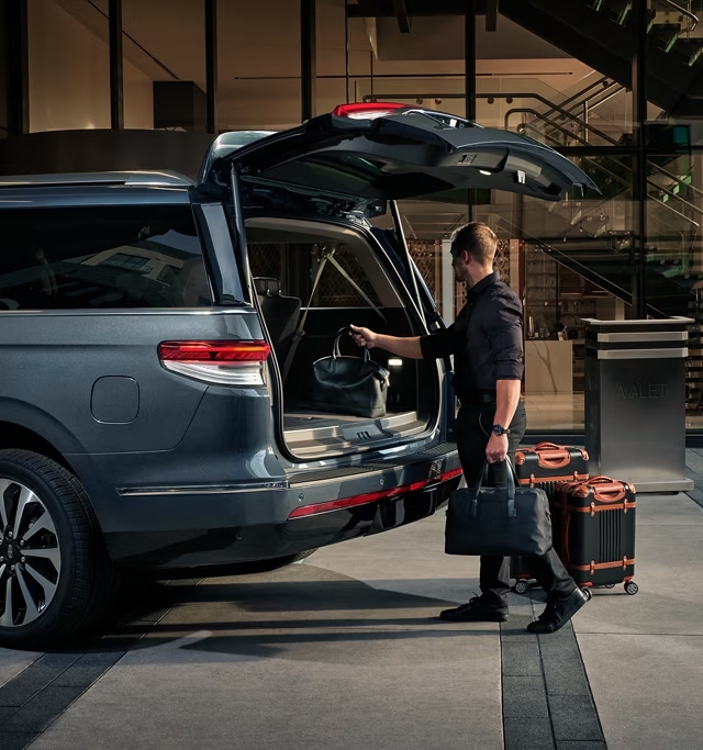 A man loads luggage into the back of a gray SUV with the trunk open. He holds a black bag in one hand and a black suitcase in the other, while two black suitcases with orange accents sit on the ground. The scene takes place at the entrance of a modern building with a sign that reads 'VALET'.