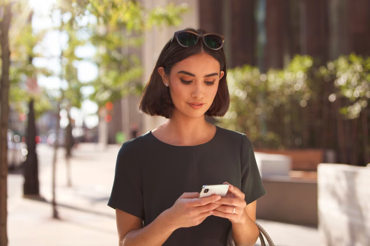 A person standing outdoors in an urban setting, interacting with a smartphone. They are wearing a dark-colored short-sleeved top with sunglasses on their head. The background features trees, buildings, and a sidewalk, illuminated by daylight.